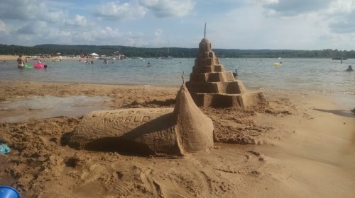 A detailed sandcastle with a pyramid shape, labeled "WORLD," on a beach with people swimming in the background.
