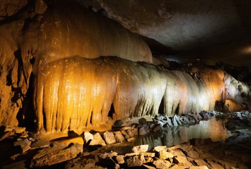 Stalactites and rock formations illuminated in a cave, reflecting in a small pool of water.
