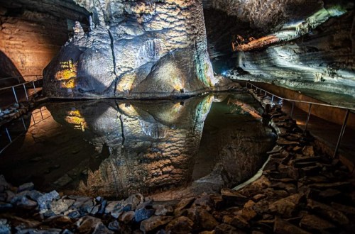 A cavern with illuminated rock formations and a still water pool reflecting the surroundings.