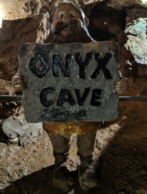 A cave figure with a beard holds a sign that reads "ONYX CAVE" in a dimly lit cave environment.