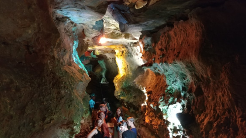 A group of people exploring a colorful cave with illuminated rock formations and a narrow passageway.