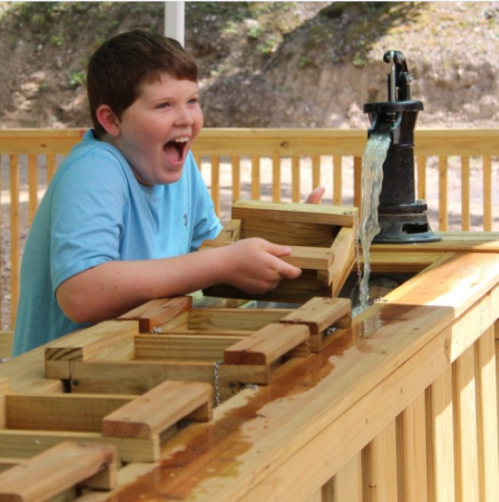 A boy joyfully plays with wooden blocks at a water feature, laughing as water flows from a pump.