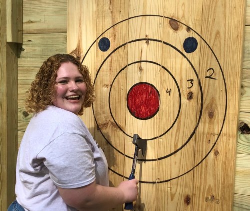 A smiling person stands next to a wooden target with concentric circles, holding an axe ready to throw.