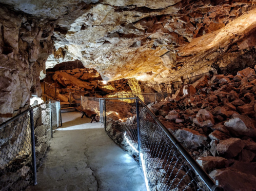 A dimly lit cave interior with rocky walls, a pathway, and a railing, showcasing natural formations and textures.