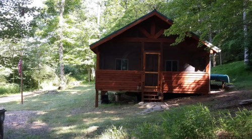 A rustic wooden cabin surrounded by trees, with a porch and grassy area in front.