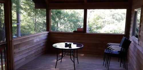 A screened porch with wooden walls, a round table, and two chairs, surrounded by greenery.