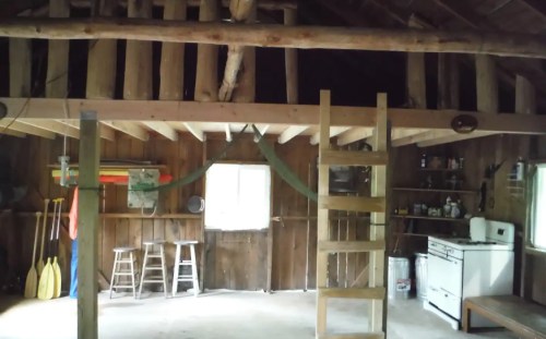 Interior of a rustic cabin featuring wooden beams, a loft, a ladder, and basic kitchen appliances.