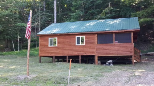 A wooden cabin with a green metal roof, surrounded by trees, featuring a flagpole with an American flag in front.