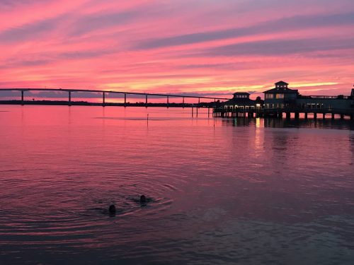 Two swimmers create ripples in the water as a vibrant sunset paints the sky in shades of pink and orange.