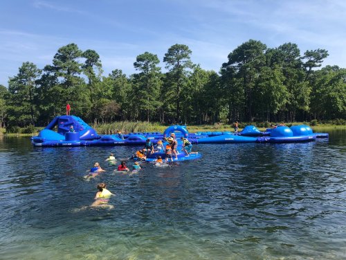 A group of children in life jackets swim near a large blue water play structure in a lake surrounded by trees.