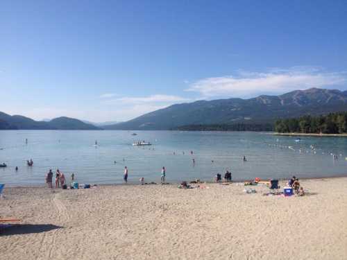 A sunny beach scene with people swimming and relaxing by a lake, surrounded by mountains and clear blue skies.