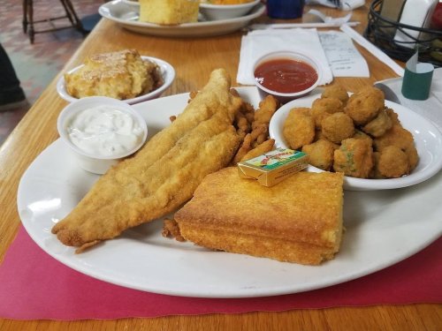 A plate of fried fish, cornbread, fried okra, and dipping sauces, served on a wooden table.