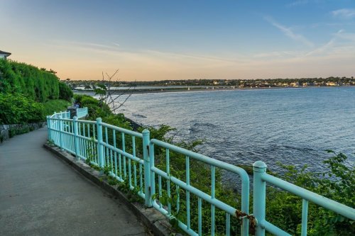 A scenic waterfront path with a turquoise railing, overlooking calm waters and a distant shoreline at sunset.