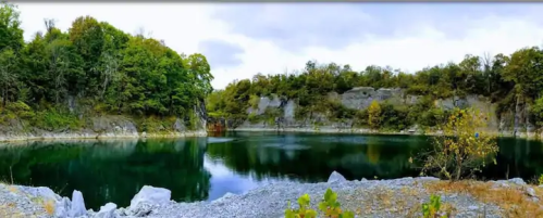 A serene quarry lake surrounded by lush greenery and rocky cliffs under a cloudy sky.