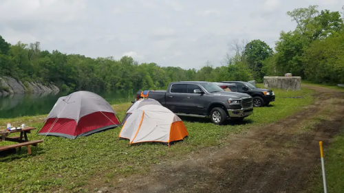A campsite by a river with two tents, picnic table, and parked trucks surrounded by greenery.