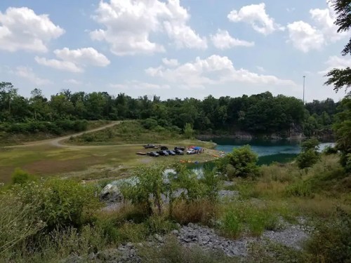 A scenic view of a quarry with a blue water body, surrounded by greenery and parked vehicles on a sunny day.