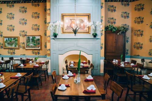 Cozy dining room with wooden tables, floral wallpaper, and a fireplace, set with white dishes and red napkins.