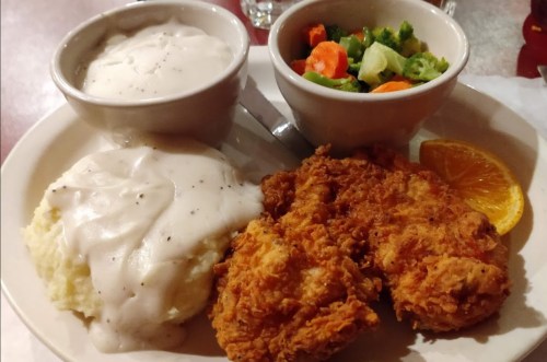 A plate with fried chicken, mashed potatoes with gravy, and a side of mixed vegetables and an orange slice.