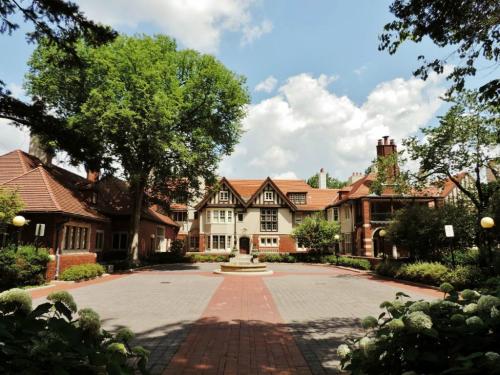 A large, elegant brick mansion surrounded by greenery and a circular driveway under a partly cloudy sky.