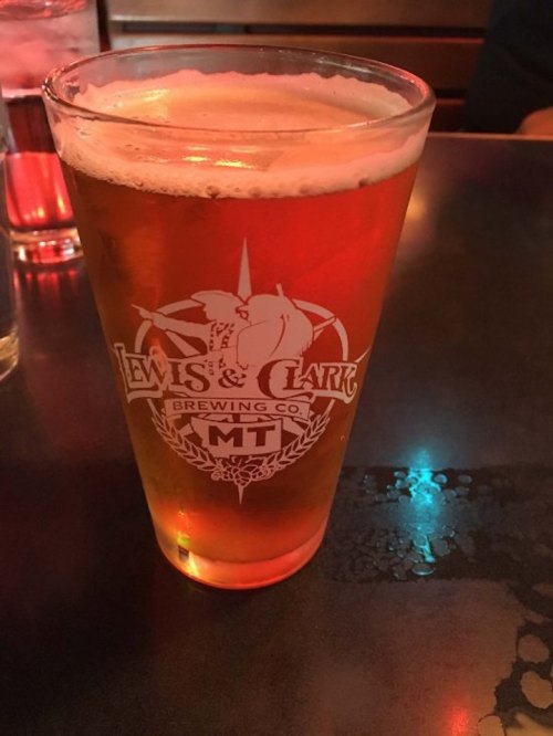 A glass of amber beer with a logo from Lewis & Clark Brewing Co. on a dark table.