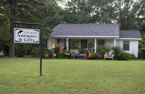 A quaint house with a sign reading "As The Crow Flies Antiques & Gifts" surrounded by greenery and colorful decorations.