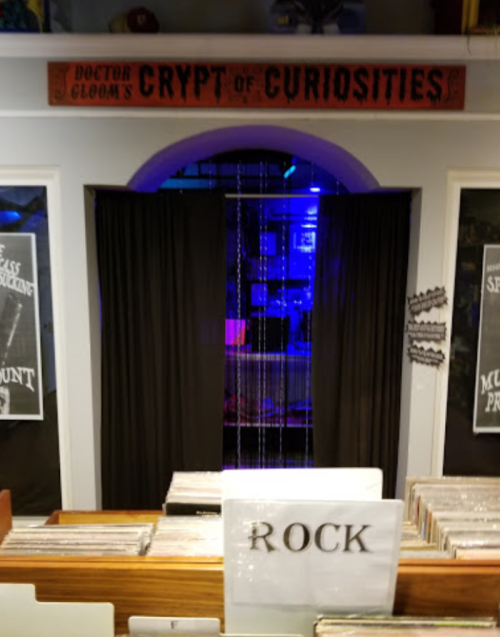 Entrance to "Doctor Gloom's Crypt of Curiosities," featuring black curtains and a sign above, with a record display in front.