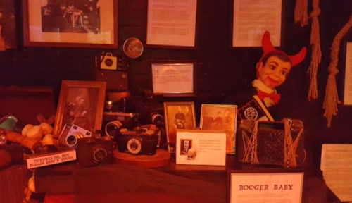 A display featuring vintage cameras, framed photos, and a puppet with devil horns in a dimly lit setting.
