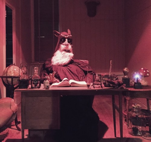 A mysterious figure with a long beard and glasses sits at a desk surrounded by potions and strange objects in a dimly lit room.