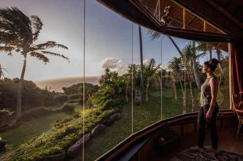 A woman stands by a large window, gazing at a sunset over the ocean, surrounded by lush greenery and palm trees.