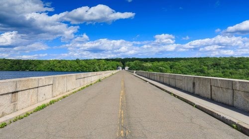 A wide, empty road stretches across a dam, flanked by lush greenery and a blue sky with fluffy clouds.