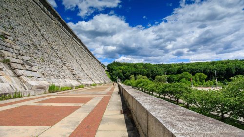 A stone dam with a sloped wall, surrounded by greenery and under a blue sky with fluffy clouds.