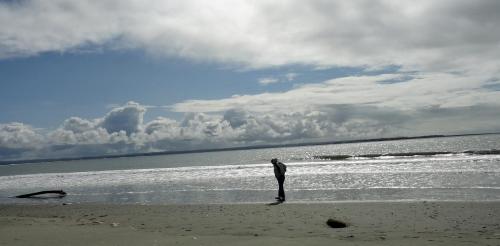 A person stands on a beach, gazing down at the sand with waves and clouds in the background.