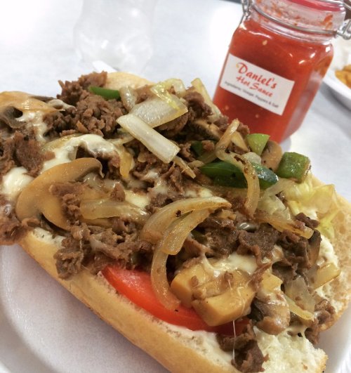 A loaded sandwich with beef, sautéed onions, mushrooms, and peppers, served with a jar of hot sauce in the background.