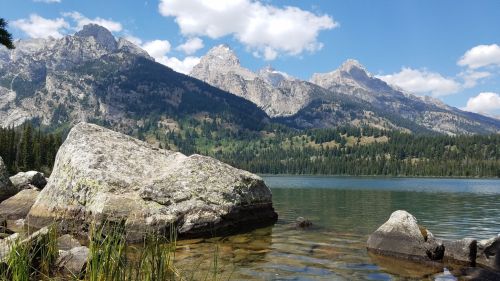 A serene lake surrounded by mountains, with clear water and rocky shores under a blue sky with fluffy clouds.