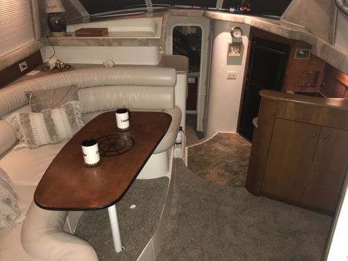 Interior of a cozy boat cabin featuring a seating area, table, and kitchenette, with soft lighting and carpeted floor.