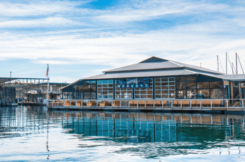 A waterfront building with large windows, reflecting in calm water, under a blue sky with scattered clouds.