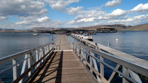 A wooden pier extends over calm water, leading to boats and a marina under a partly cloudy sky.