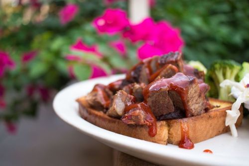 A plate of grilled meat on toast, drizzled with sauce, accompanied by broccoli and salad, with pink flowers in the background.