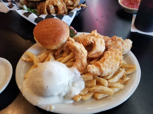 A plate with fried chicken strips, French fries, a biscuit, and gravy on top of the fries.