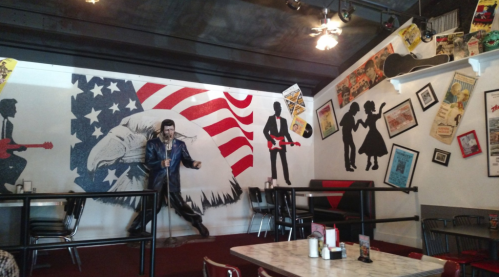 A vibrant diner interior featuring a mural of a singer, American flag, and vintage music-themed decor on the walls.
