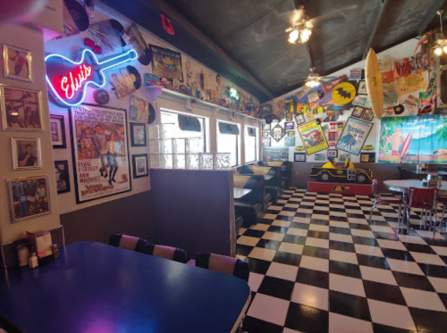 A retro diner with checkered floors, colorful wall decor, and neon signs, featuring booths and a vibrant atmosphere.
