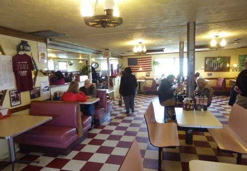 A cozy diner interior with checkered floors, booths, and patrons enjoying meals, decorated with an American flag.
