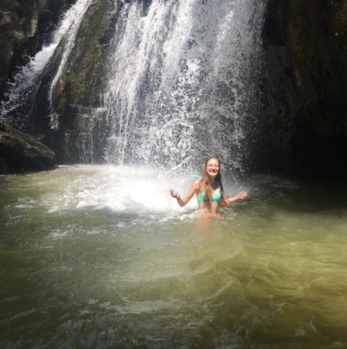 A girl in a bikini stands in a river, smiling in front of a waterfall surrounded by rocks and greenery.