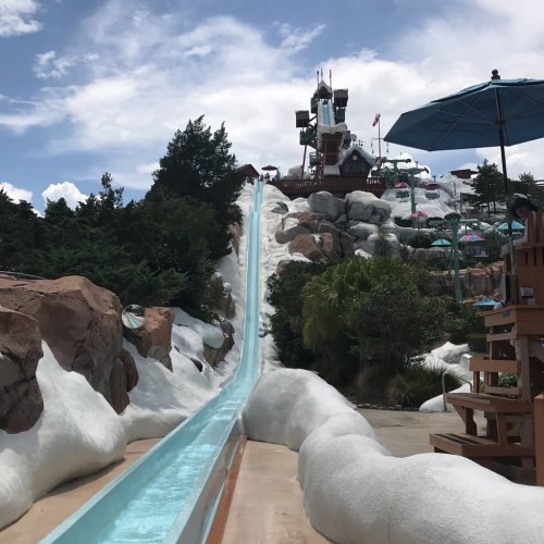 A tall water slide descends from a rocky structure, surrounded by trees and umbrellas under a blue sky.