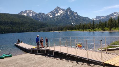 A scenic lake surrounded by mountains, with people on a dock and kayaks nearby under a clear blue sky.