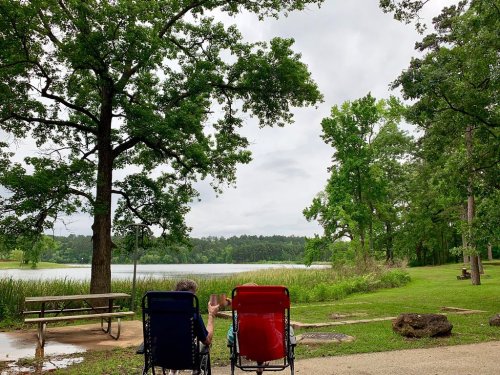 Two people sit in red and blue chairs by a lake, surrounded by trees under a cloudy sky.