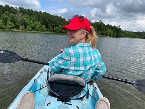 A woman in a red cap and plaid shirt smiles while kayaking on a calm lake surrounded by trees.