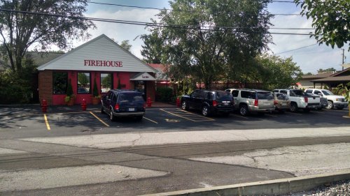 A parking lot with several vehicles in front of a building labeled "Firehouse" surrounded by trees.