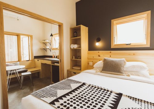 Cozy bedroom with a wooden bed, patterned blanket, and a view into a bright kitchen area with natural light.