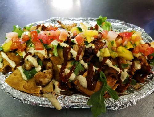 A platter of nachos topped with grilled chicken, fresh salsa, drizzled sauces, and garnished with arugula.